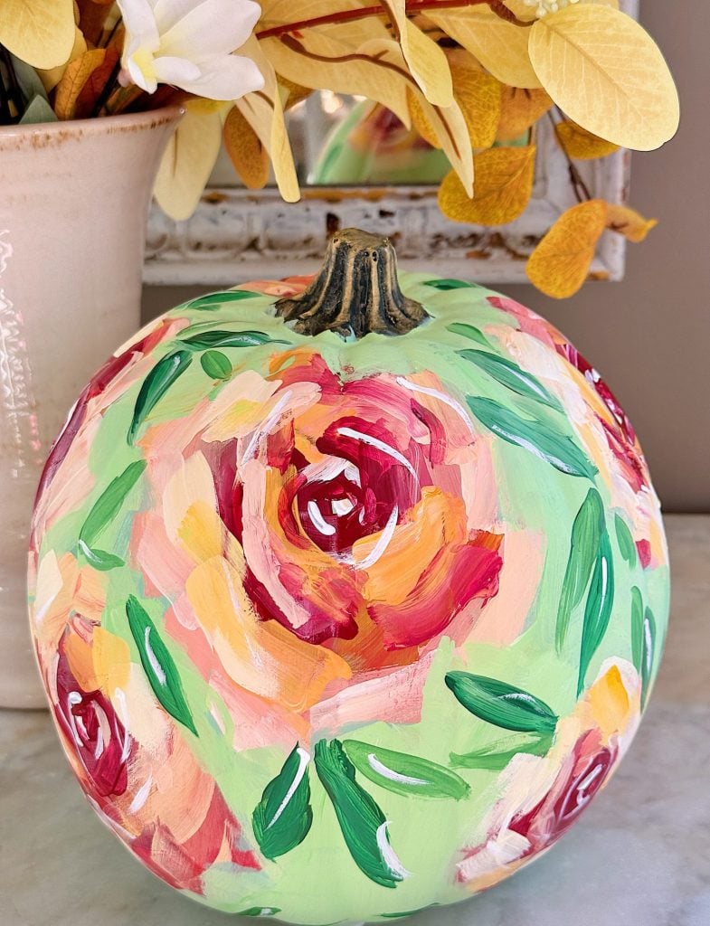 A pumpkin painted with colorful floral designs sits on a table next to a potted plant with yellow leaves.