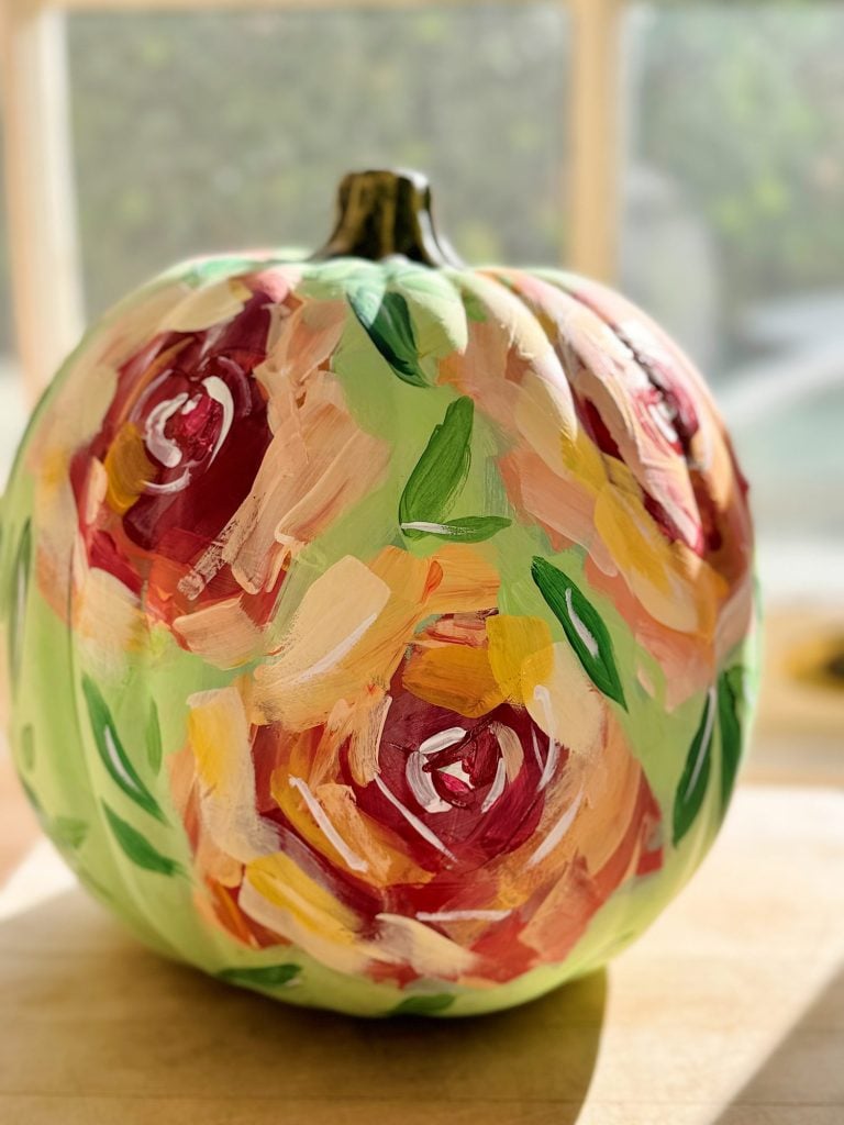 A pumpkin painted with colorful floral designs in red, pink, and green, illuminated by natural light.