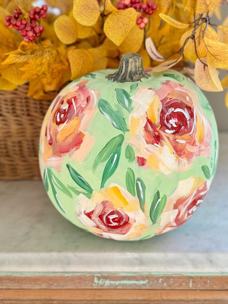 A pumpkin painted with colorful floral designs is placed in front of autumn leaves in a woven basket.