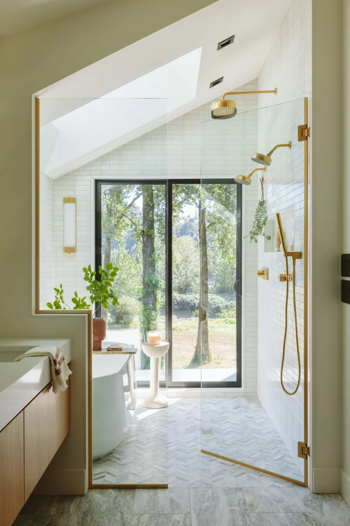 Modern bathroom with glass shower, gold fixtures, indoor plants, large window with forest view, and geometric tile flooring.