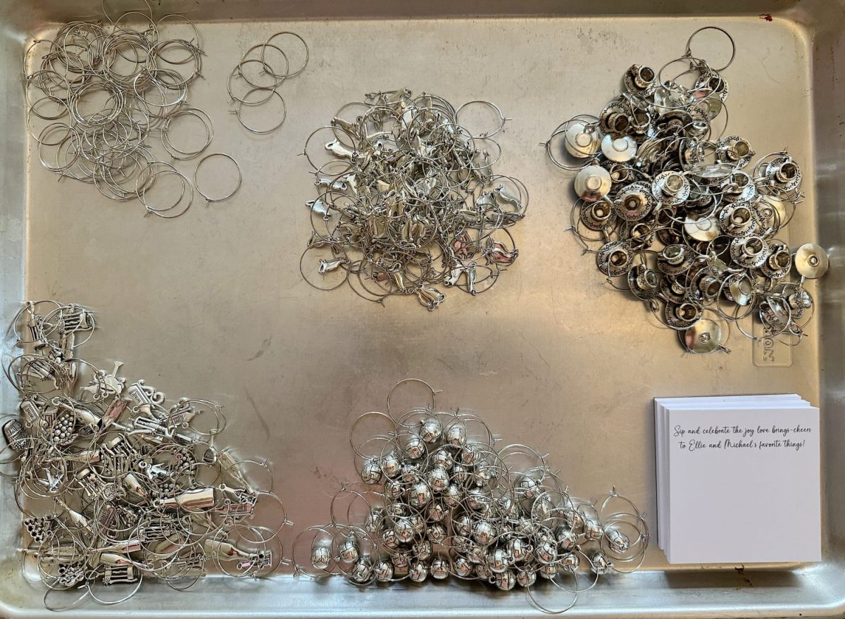A tray displays sorted metal wine glass charms in various designs, with a small white note card in the bottom right corner.