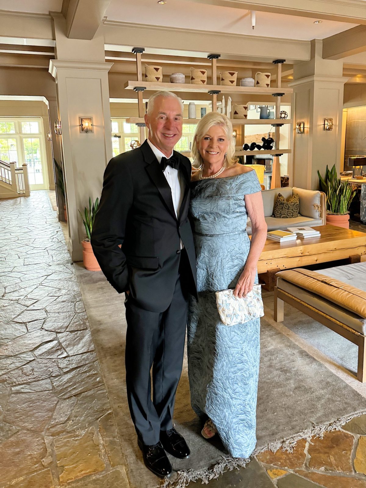 A couple dressed in formal attire stands together in a stylish interior space with modern decor. The man is in a suit, and the woman is in a blue gown.
