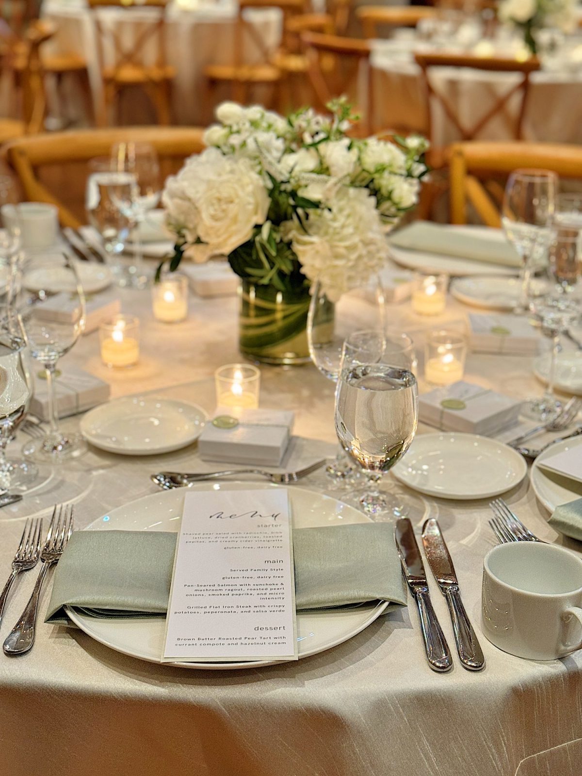 Elegant table setting with white floral centerpieces, candles, and neatly arranged cutlery around a plate with a menu.