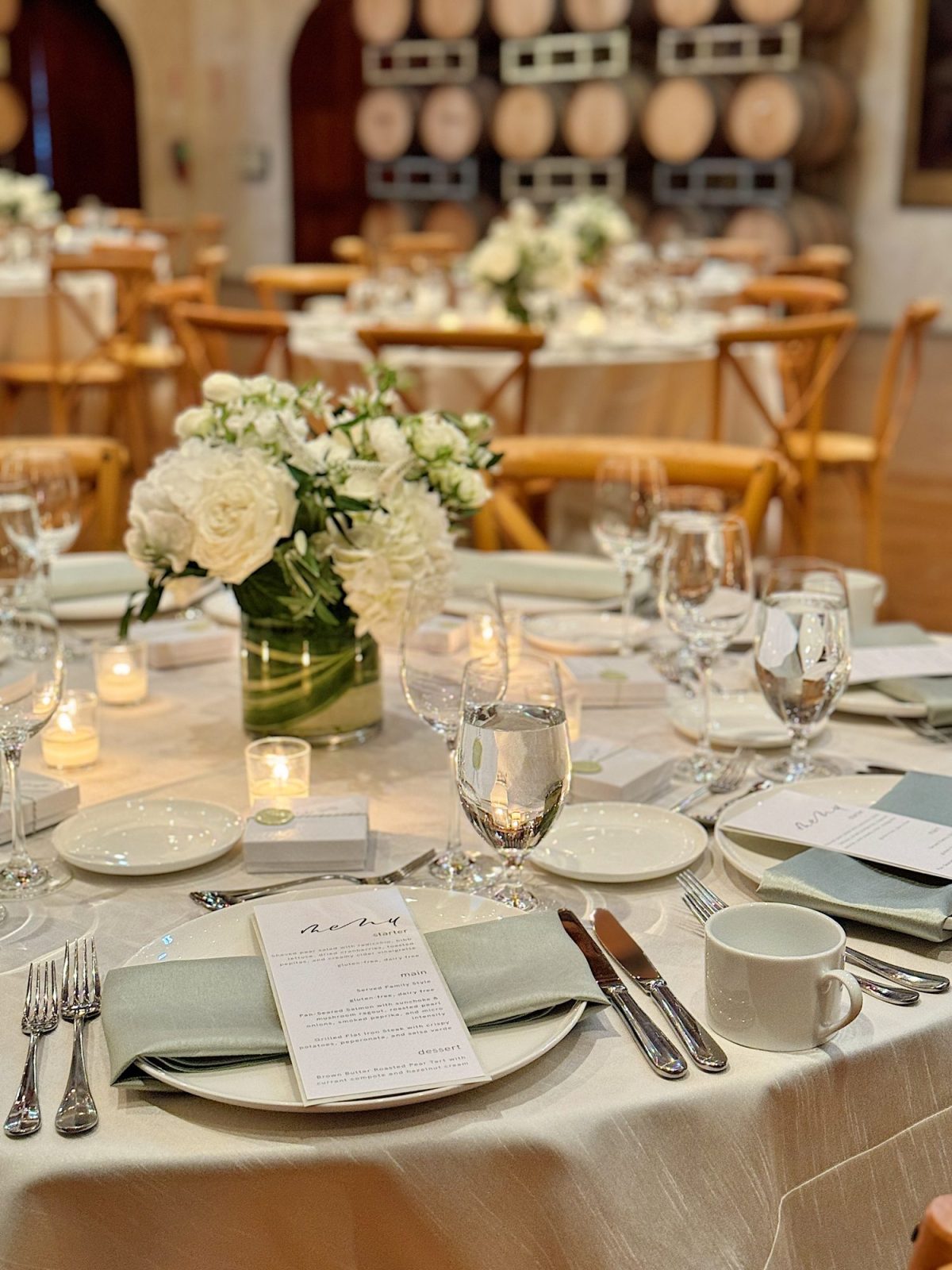 Elegant table setting with white floral centerpiece, menus on plates, wine glasses, and candles. Wooden chairs and stacked barrels in the background.