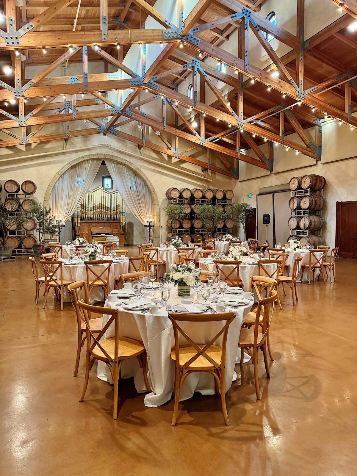 Rustic banquet hall with wooden beams, round tables set with white tablecloths, elegant place settings, and floral centerpieces. Wine barrels are stacked against the walls.