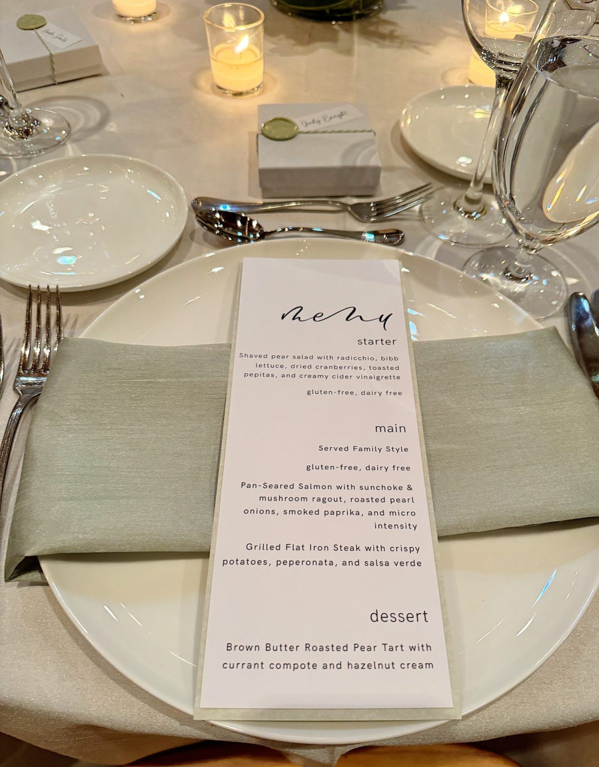 A formal dining table setting with a menu on a white plate. The menu lists starter, main, and dessert options, including a pear salad, salmon, and a pear tart.