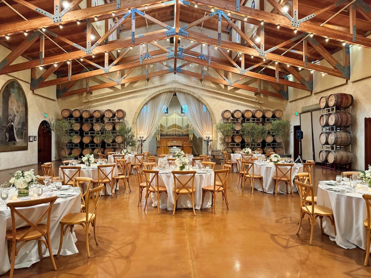 Rustic event hall with wooden beams, round tables set with white cloths, and beige chairs. Wine barrels line the walls, and a large archway is curtained at the back.