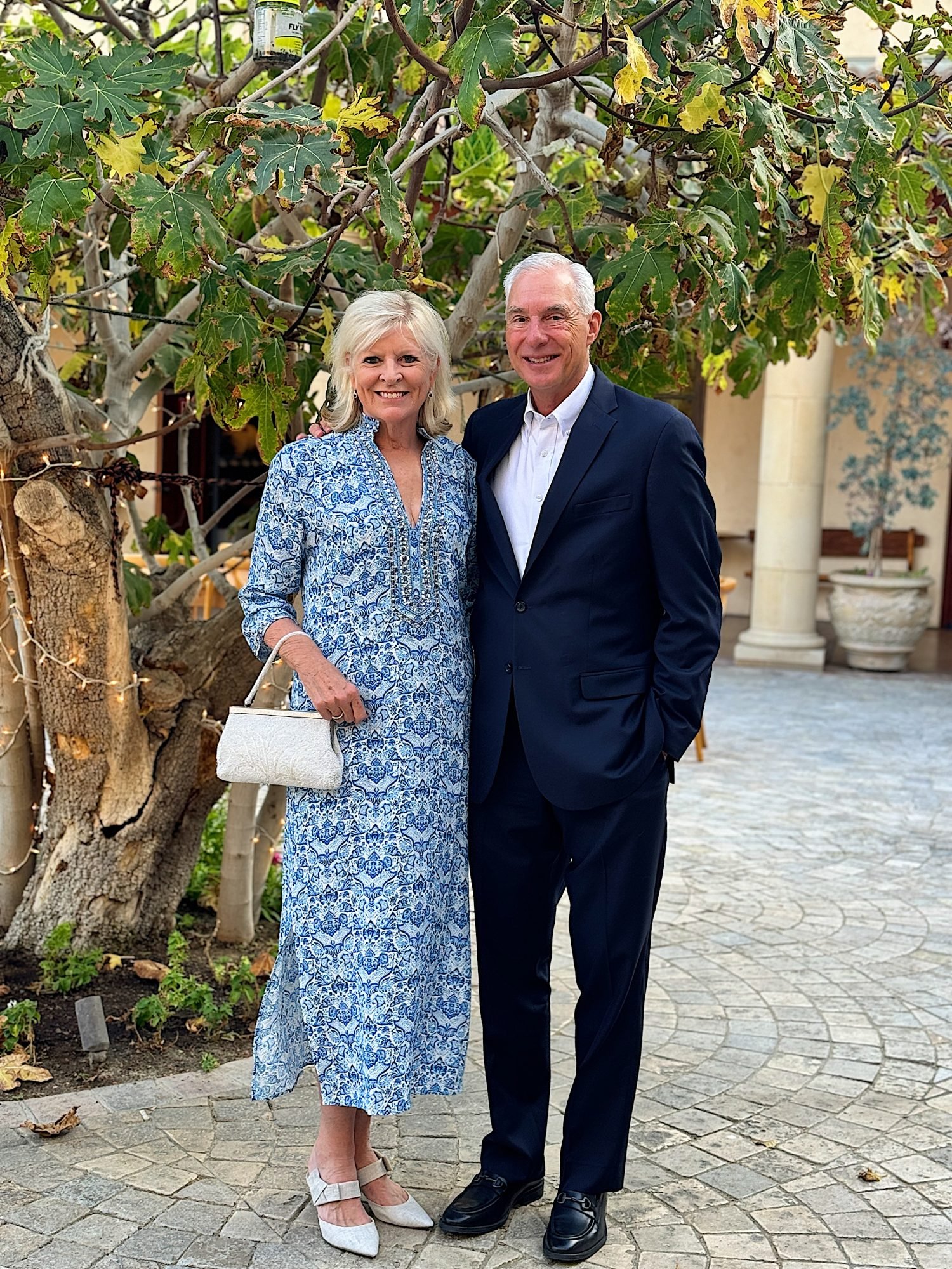 A woman in a blue patterned dress and white shoes stands next to a man in a dark suit. They are outdoors on a paved area with trees and a building in the background.