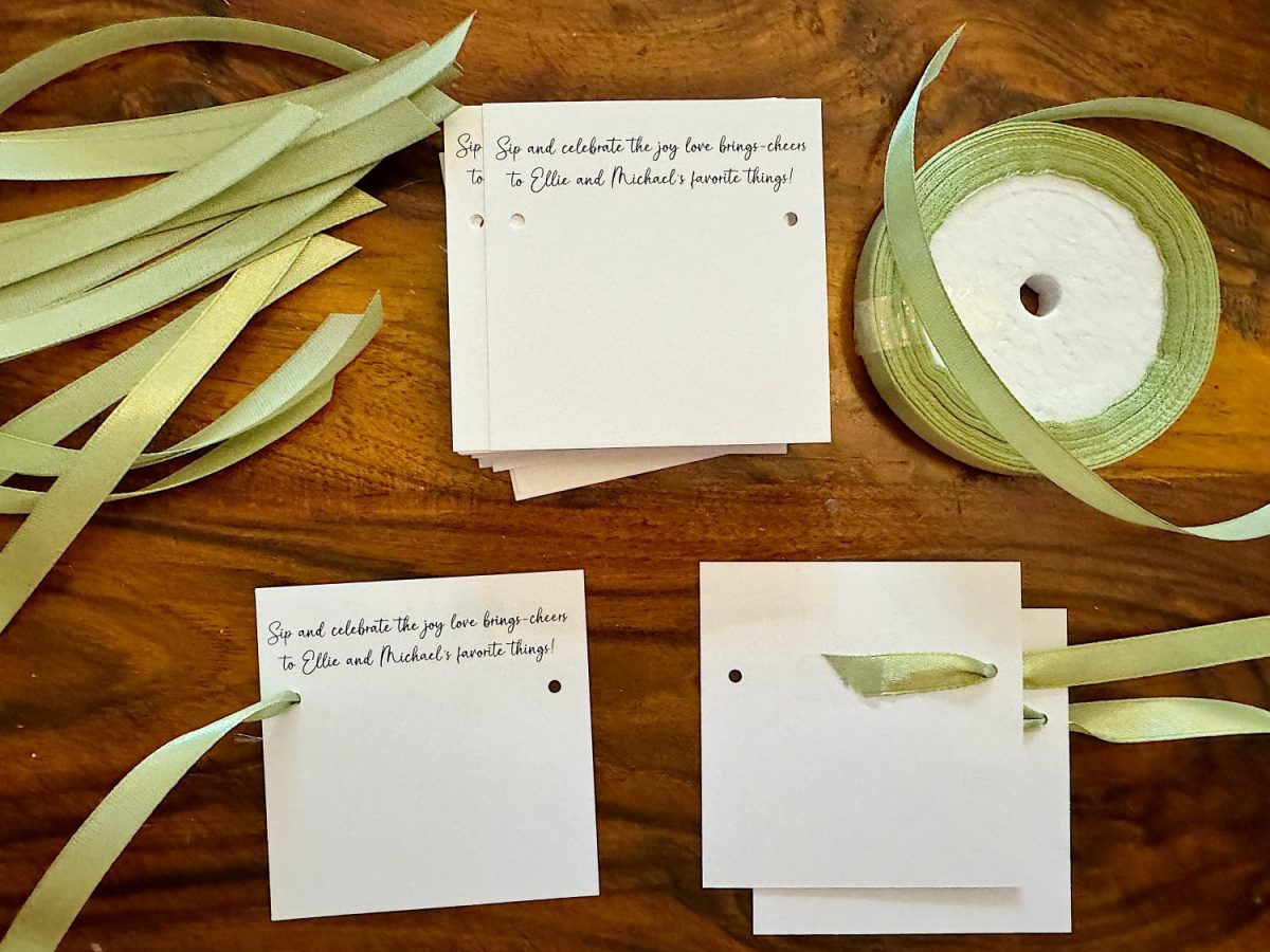 Stacks of square paper cards, each with green ribbon and text. Nearby are loose ribbons and a spool on a wooden surface.