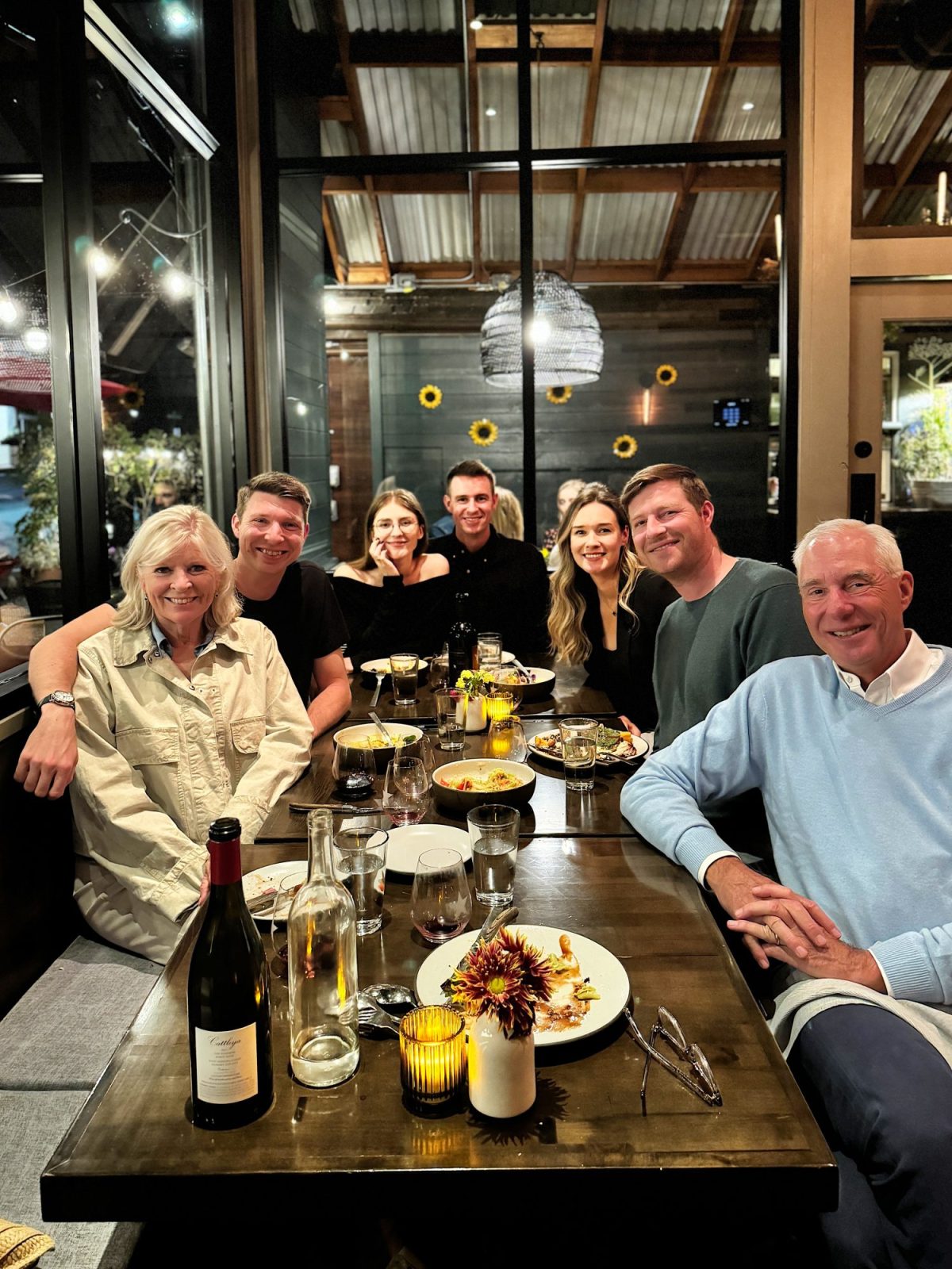 A group of seven people sitting around a dining table in a restaurant, with plates and glasses, smiling at the camera.