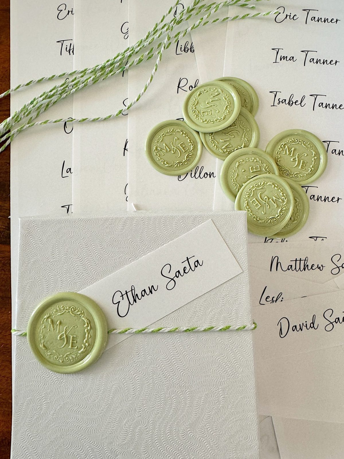 A stack of white invitations with green wax seals and twine. A card with "Ethan Saeta" is visible. Wax seals have initials "MS" and "JE.