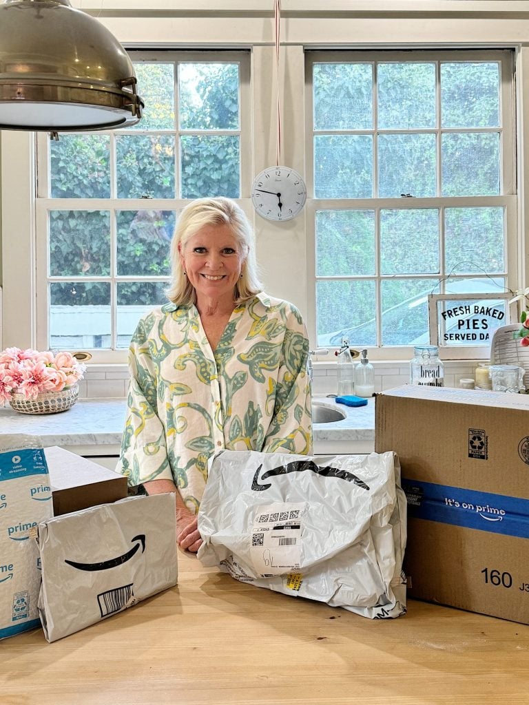 A person stands in a kitchen surrounded by Amazon packages on a wooden table. A window, clock, and flowers are in the background.