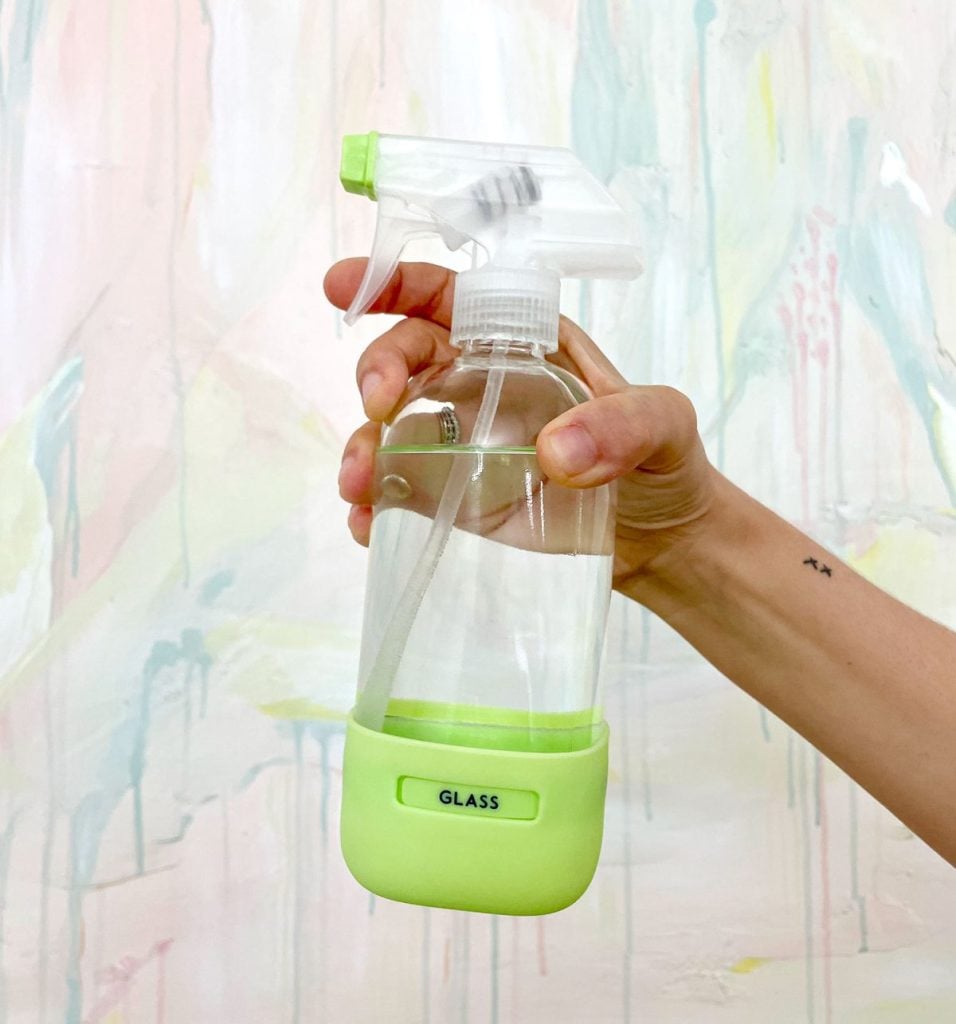 A hand holding a clear spray bottle with a green base and a label that reads "GLASS" against a pastel-colored wall.