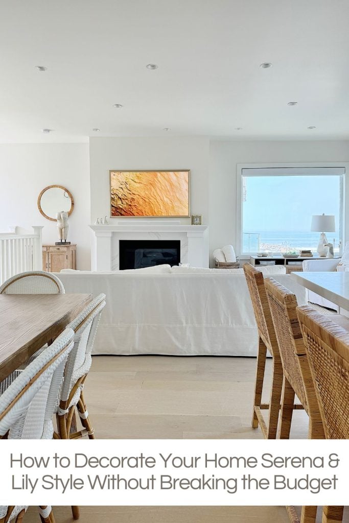 A bright, modern living room with white furniture, a large painting above the fireplace, and wicker chairs around a dining table. Large windows offer a scenic view.