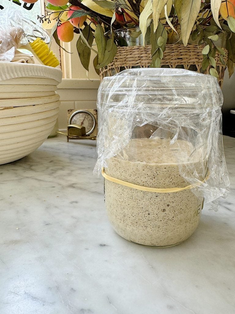 A glass jar with sourdough starter covered in plastic wrap, secured by a rubber band.