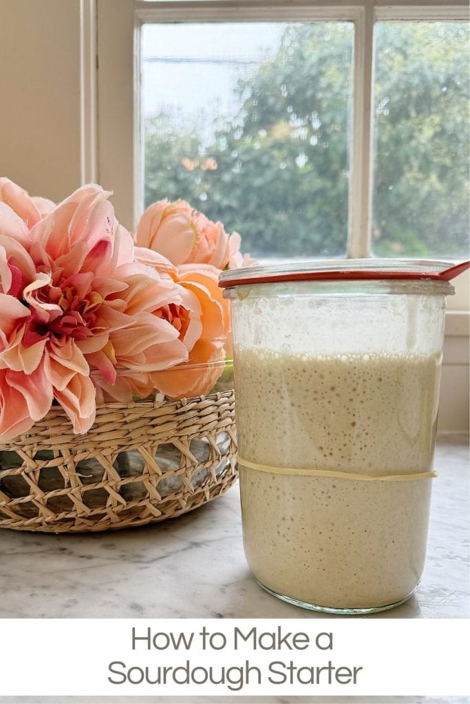 Jar of sourdough starter beside a basket of pink flowers on a marble counter, with a window in the background.