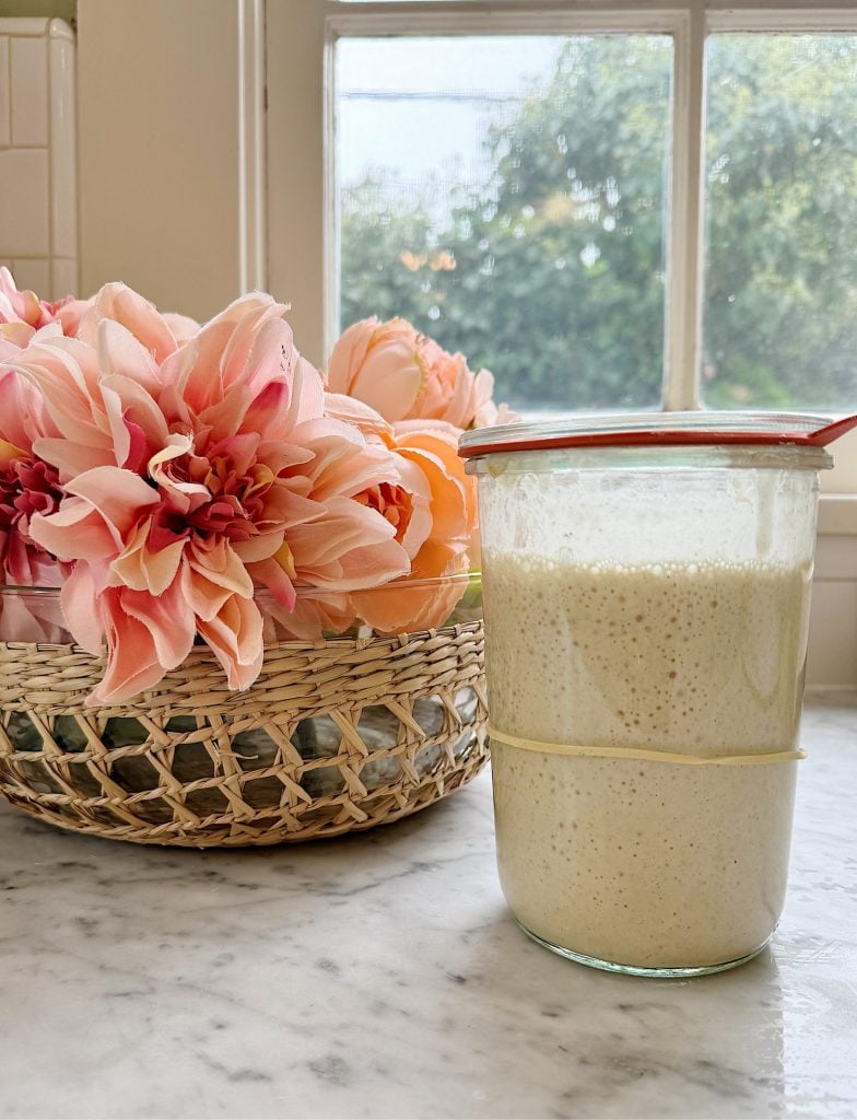 Glass container with sourdough starter and a glass lid with flowers nearby.