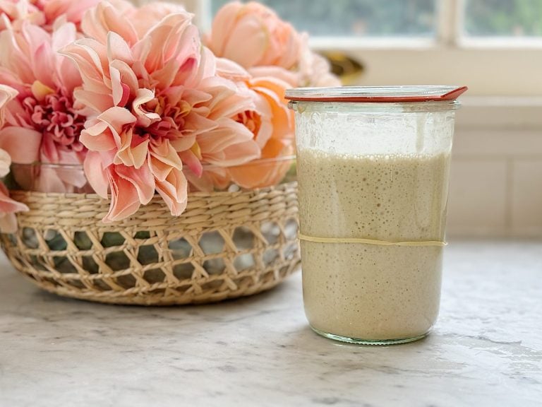 Glass container with sourdough starter and a glass lid with flowers nearby.