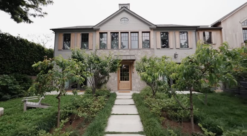 A two-story house with a stone facade, large windows, and a wooden front door is surrounded by a lush garden with small trees and a pathway leading to the entrance.