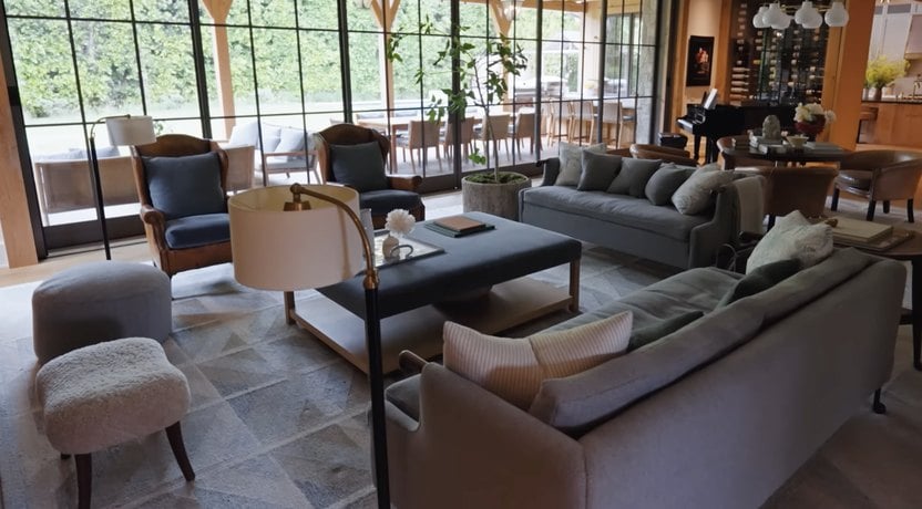 Spacious living room with large windows, two sofas, armchairs, a coffee table, and a lamp. The room opens to an outdoor patio area with a dining table and chairs. Plants and modern decor are visible.