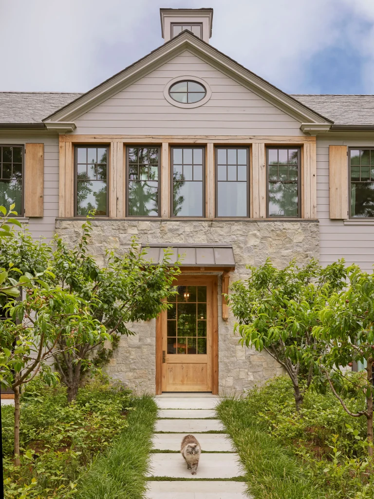 A stone and wood house with tall windows, a wooden door, and a lush front garden. A cat walks on the stone path leading to the entrance.