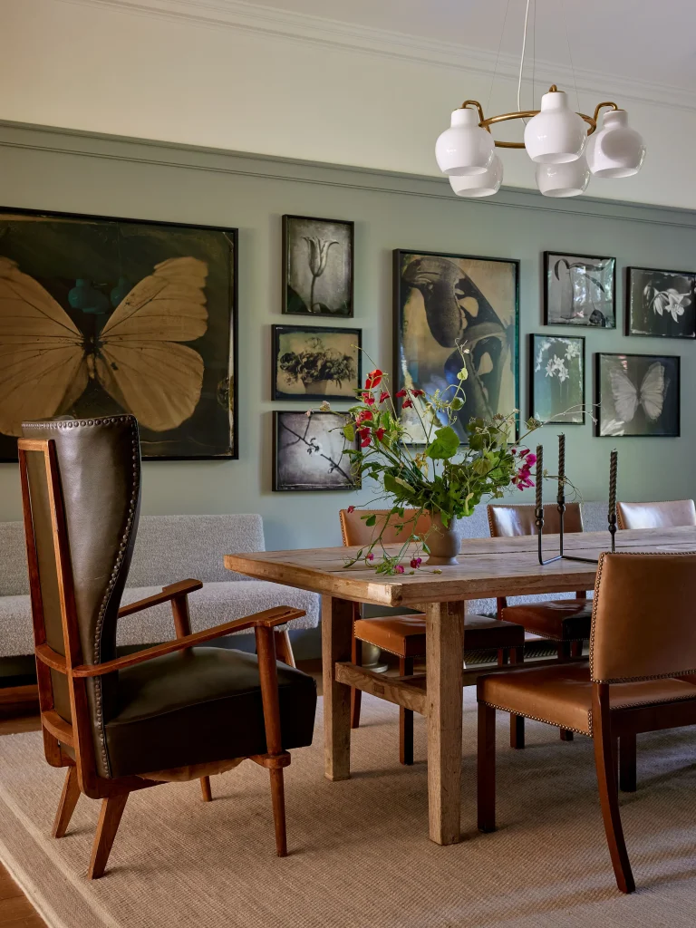 Dining room with a wooden table, leather chairs, a floral centerpiece, and botanical art on the walls. A modern chandelier hangs above the table.