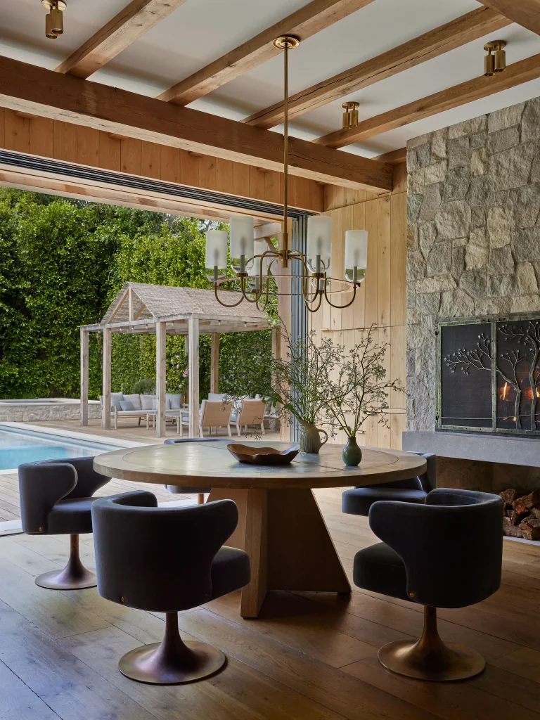 Dining area with a round wooden table and four black chairs, chandelier overhead, large glass doors open to an outdoor patio with a pool, lounge chairs, and a pergola, stone fireplace visible.