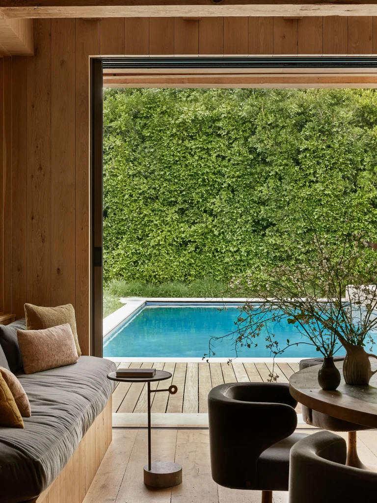 A cozy wooden interior opens to a poolside view, featuring a seating area with a sofa, chairs, table, and decorative plants near a large window.