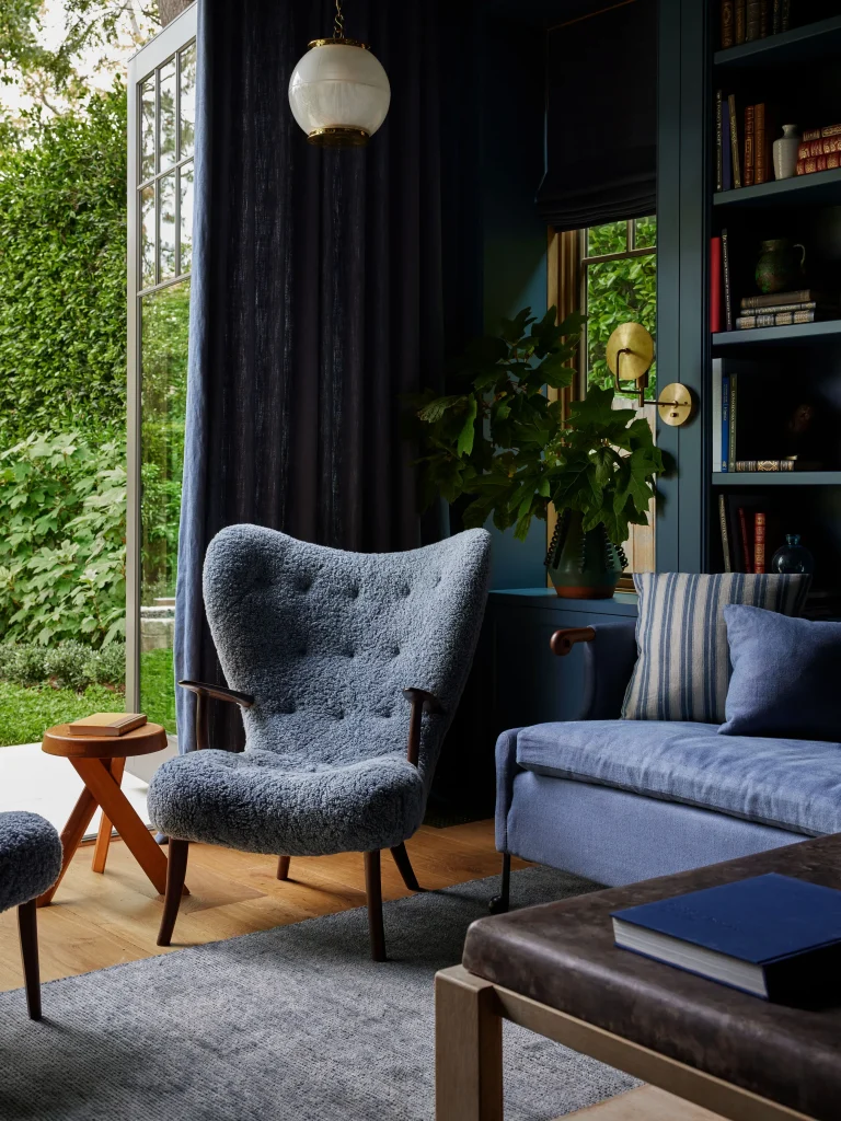 A cozy living room features a blue armchair, matching sofa, small wooden table, bookshelf, and large window overlooking greenery.