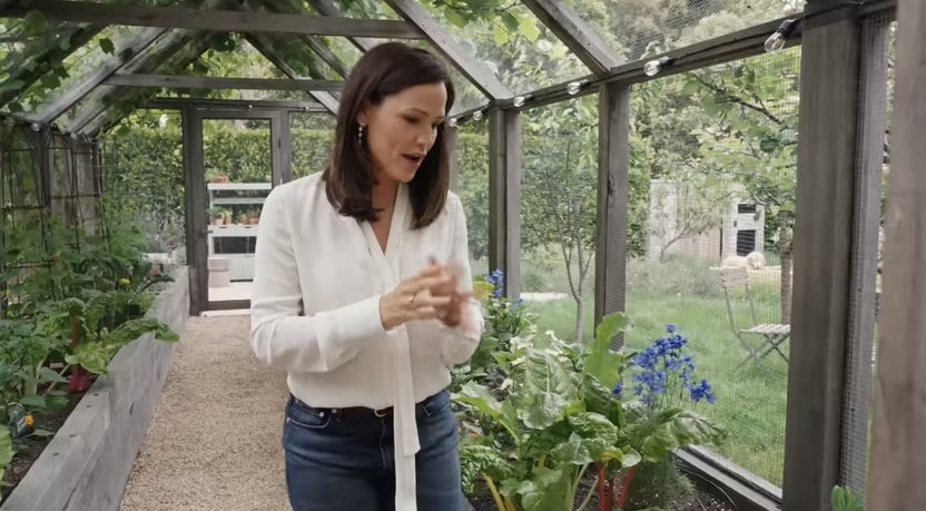 A person in a white blouse and jeans stands inside a greenhouse with plants on both sides, discussing or explaining something.