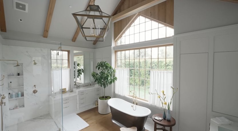 A modern bathroom features a freestanding bathtub, large windows with white curtains, a glass shower, white cabinetry, potted plant, and wooden ceiling beams.