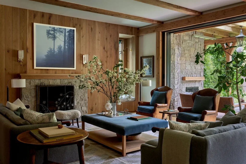 A cozy living room with wooden walls, a stone fireplace, two cushioned grey sofas, armchairs, a large coffee table with a floral arrangement, and an open view to a stone patio with greenery.