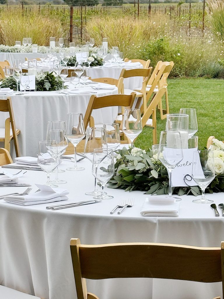 Outdoor wedding reception setup with round tables covered in white cloths, wooden chairs, and table centerpieces of greenery. Each setting includes wine glasses and folded napkins.