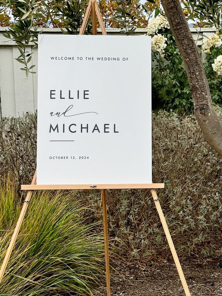 Wedding sign on an easel reads, "Welcome to the wedding of Ellie and Michael, October 12, 2024," set outdoors with greenery and flowers.