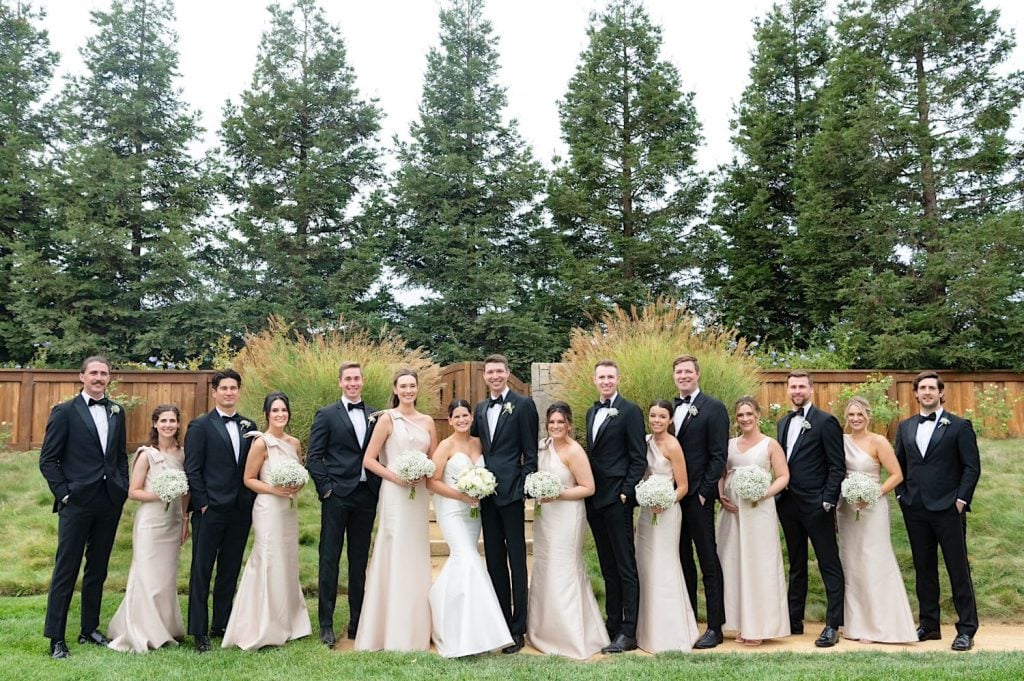 A wedding party poses outdoors, with the couple in the center surrounded by bridesmaids in champagne dresses and groomsmen in black suits, against a backdrop of tall trees and a wooden fence.