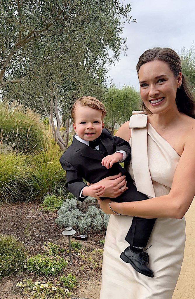 A woman in a white dress holds a young boy in a black suit, standing on a garden path with greenery in the background.