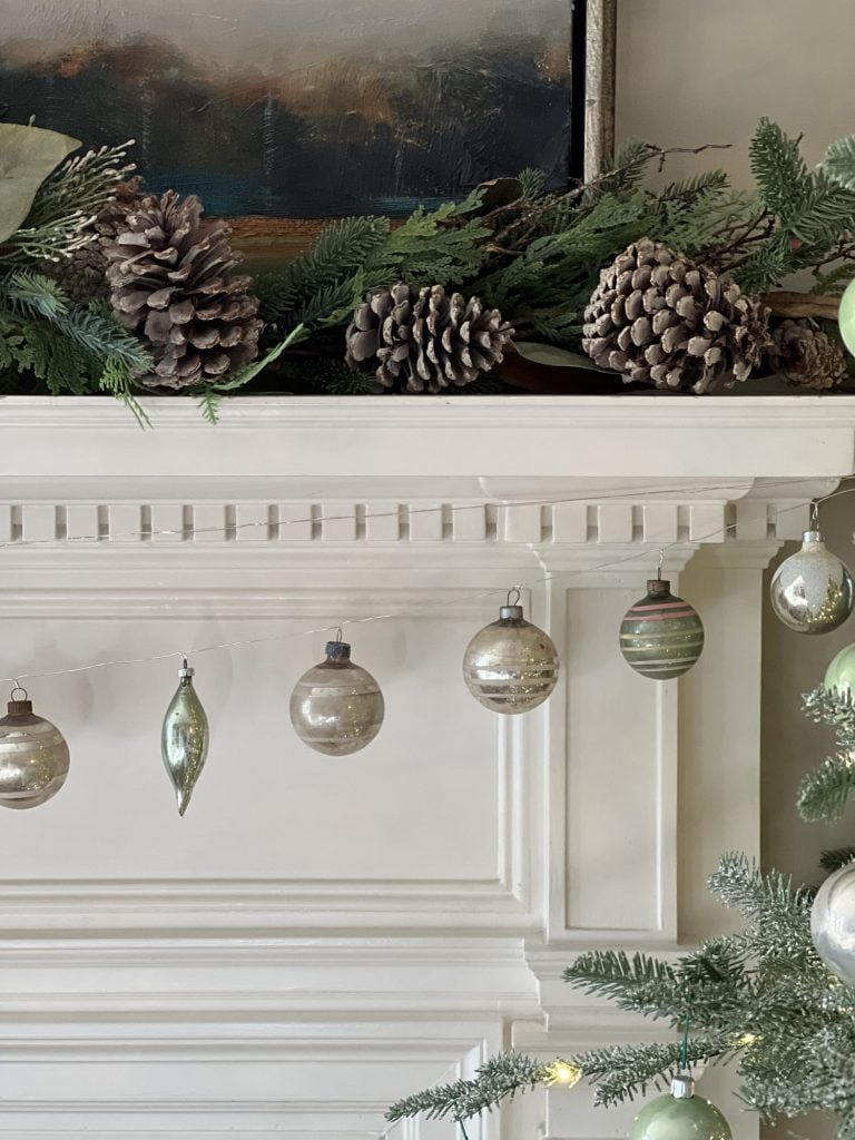 A fireplace mantel decorated with pinecones, evergreen branches, and a row of silver and green ornaments hanging below. A partial view of a decorated Christmas tree is visible on the right.