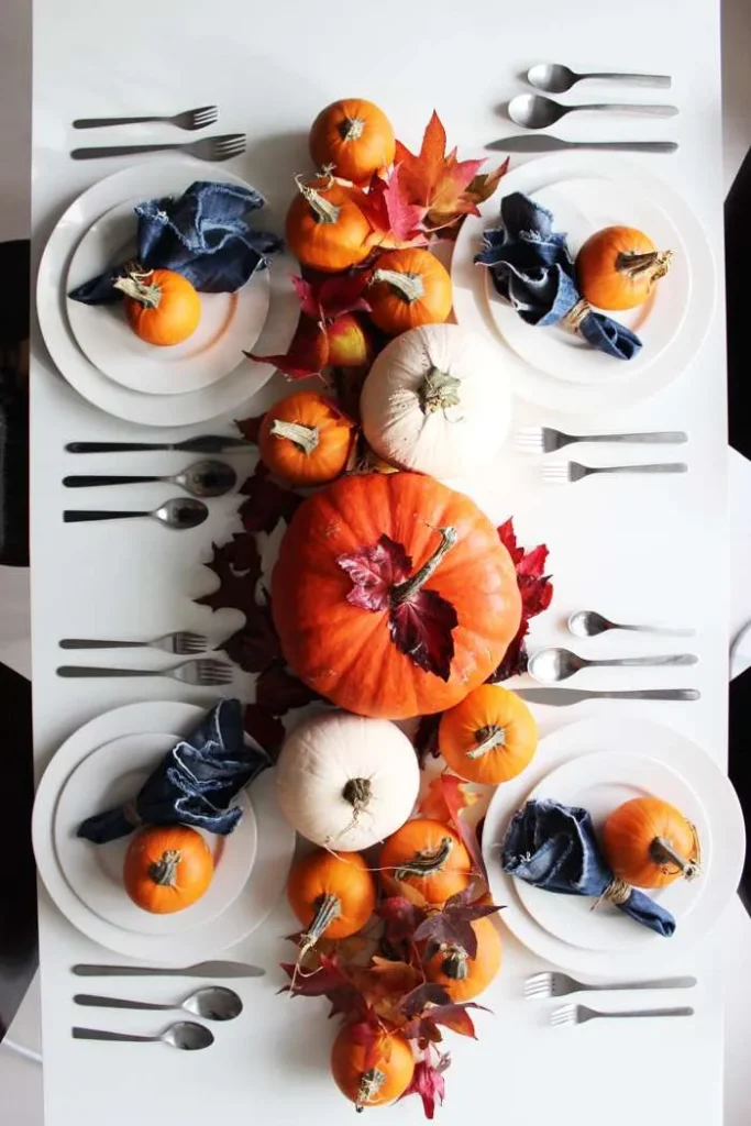 A table set with plates, silverware, and napkins features an autumn centerpiece of pumpkins and fall leaves.