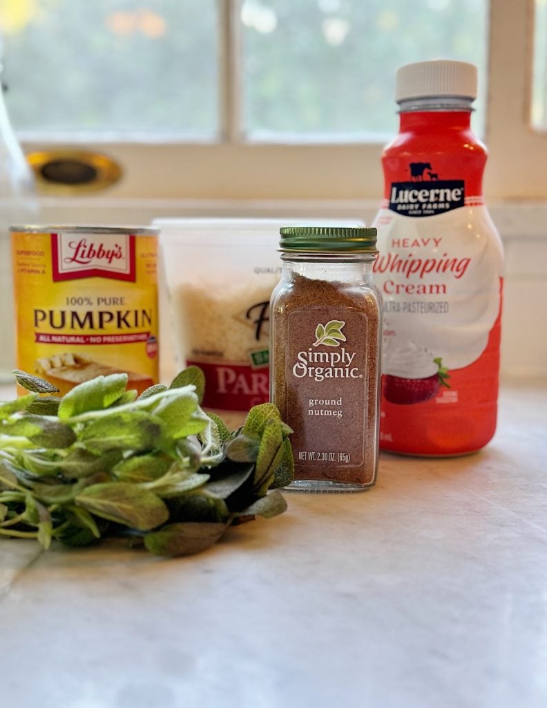 Various cooking ingredients on a countertop: fresh herbs, ground nutmeg, Parmesan cheese, a can of pumpkin, and a bottle of heavy whipping cream.