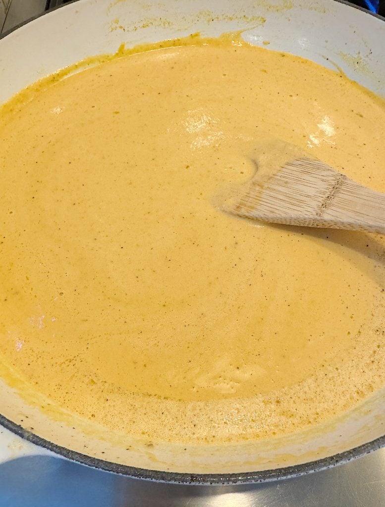 A wooden spoon rests in a large pan filled with creamy orange sauce.