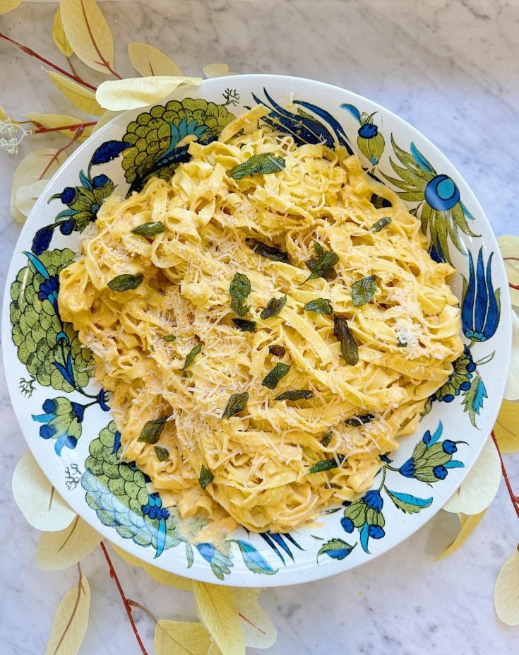 A bowl of creamy pasta topped with grated cheese and herbs on a decorative blue and green floral plate, placed on a marble surface surrounded by light-colored leaves.