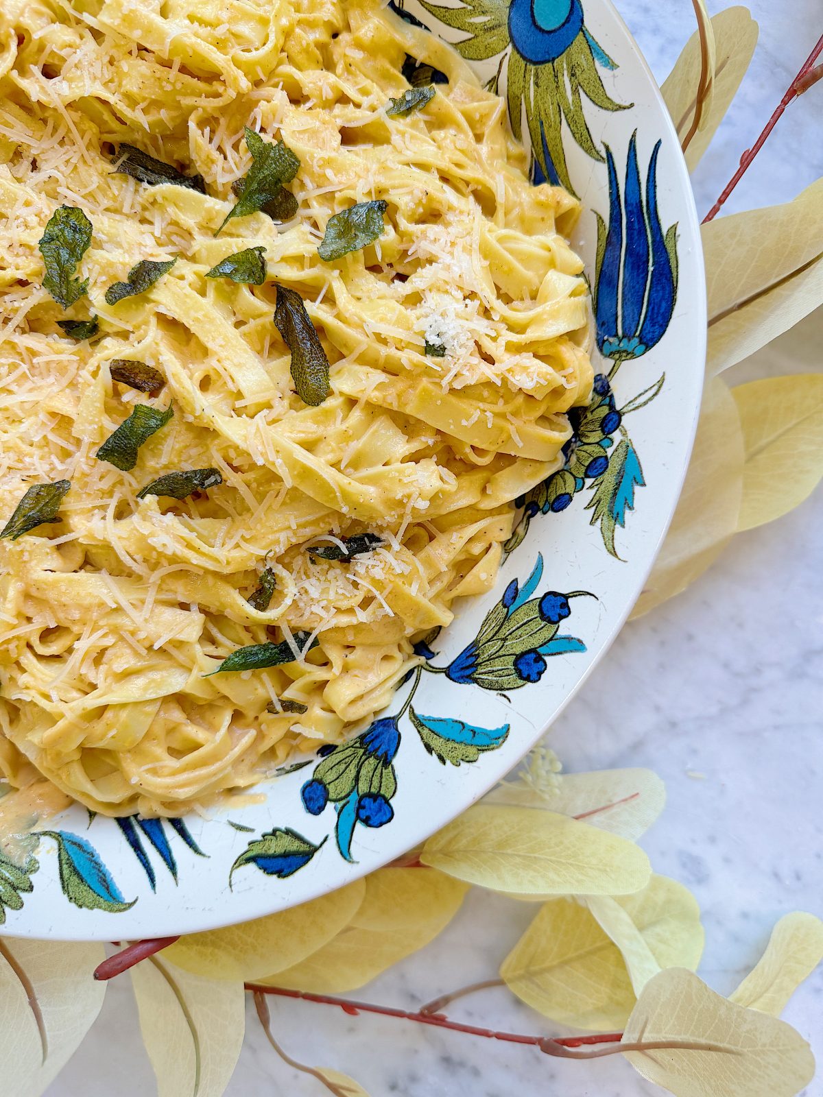 A decorative plate contains fettuccine pasta coated in creamy sauce, garnished with grated cheese and fresh herbs, set on a light-colored surface with leaf accents.