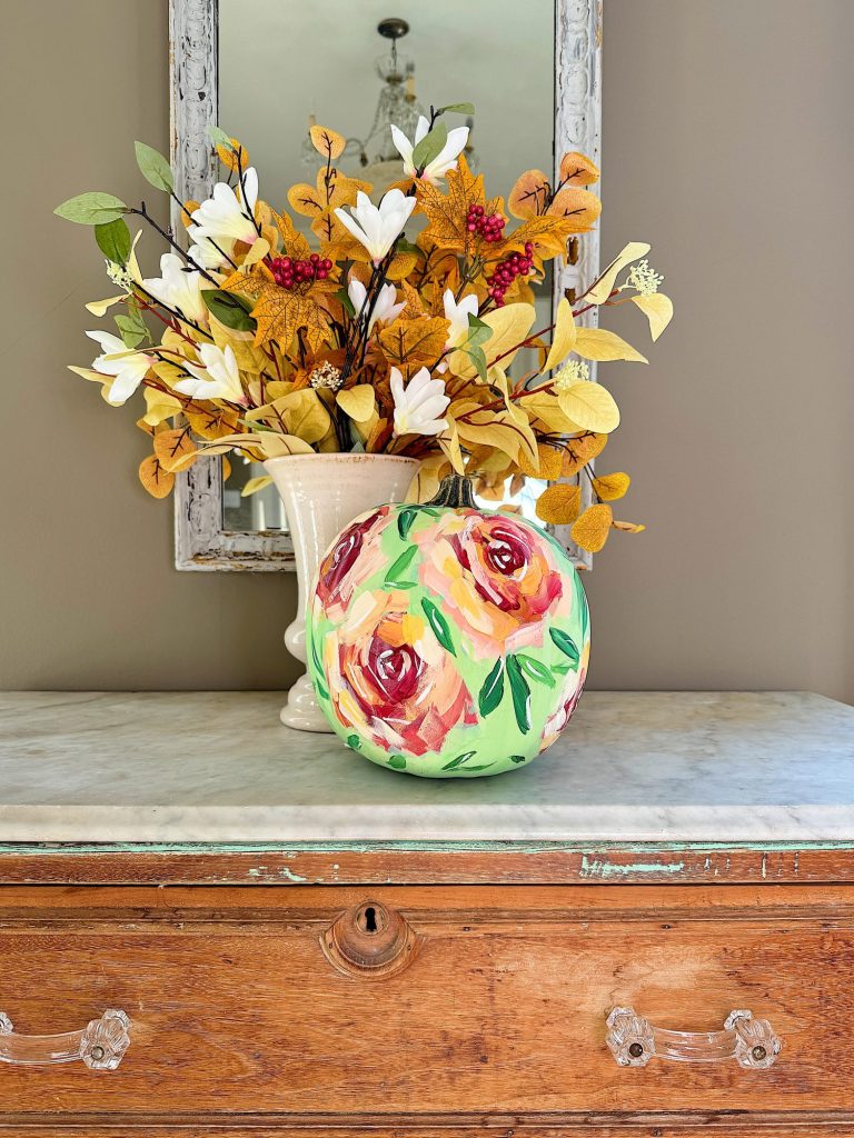 A painted pumpkin with floral designs sits on a wooden dresser, next to a vase of autumn-colored flowers, beneath a wall mirror.