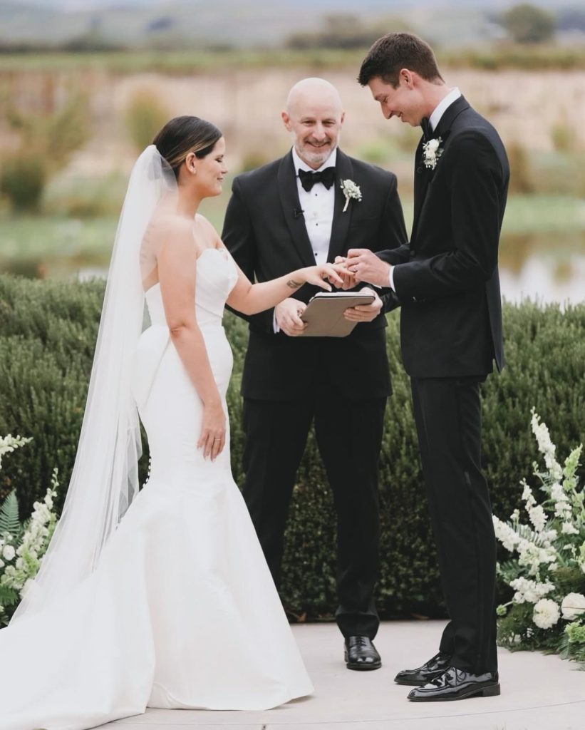 A bride and groom exchange rings during an outdoor wedding ceremony officiated by a man in a black suit.