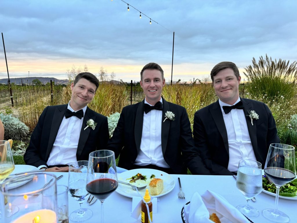 Three men in formal wear sit at an outdoor table set with glasses and plates, under strings of lights, with a scenic background of fields and a cloudy sky.