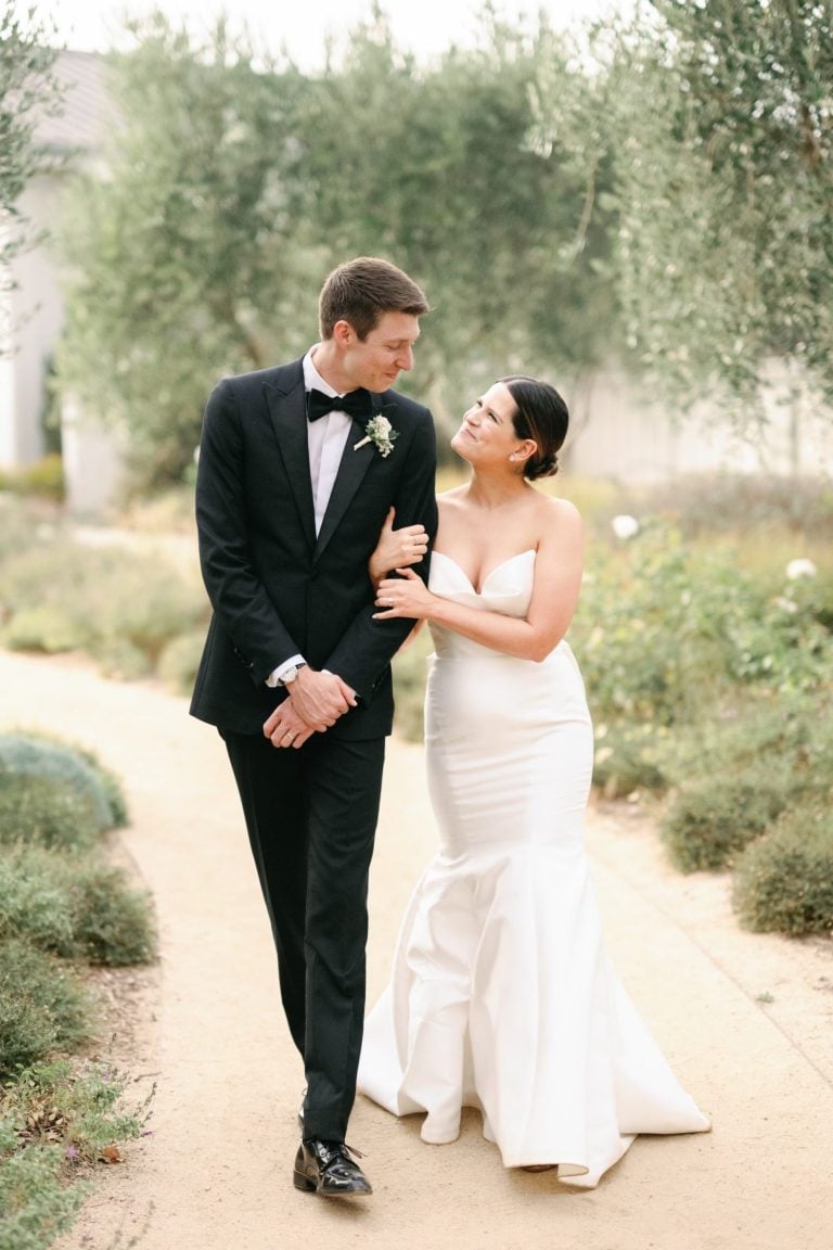 A couple walks arm in arm on a garden path, dressed in formal wedding attire.