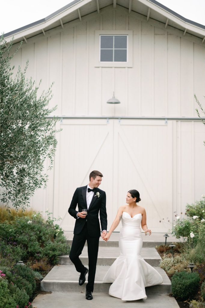 A couple in formal attire walks hand-in-hand in front of a white barn surrounded by greenery.