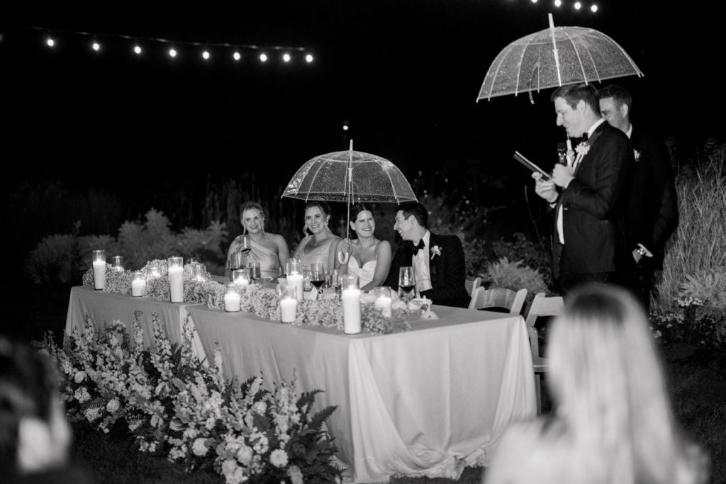 A bridal party sits at an outdoor table under umbrellas during a nighttime event.