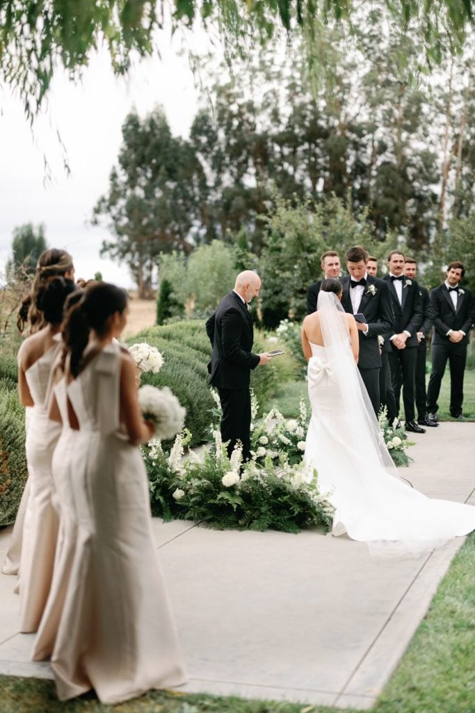 A wedding ceremony outdoors with a bride and groom facing an officiant. Bridesmaids in light dresses stand on the left, and groomsmen in black suits stand on the right.