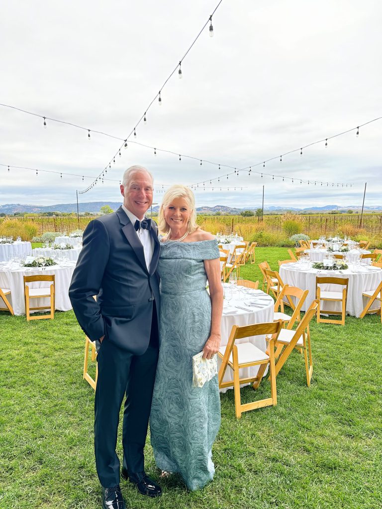 A couple dressed in formal attire stands on grass with tables and string lights in the background.